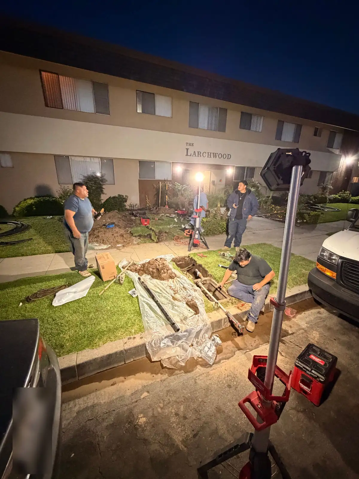 Mainline Plumbing crew replacing an apartment building sewer lateral overnight, lawn covered in protective tarps and work lights set up